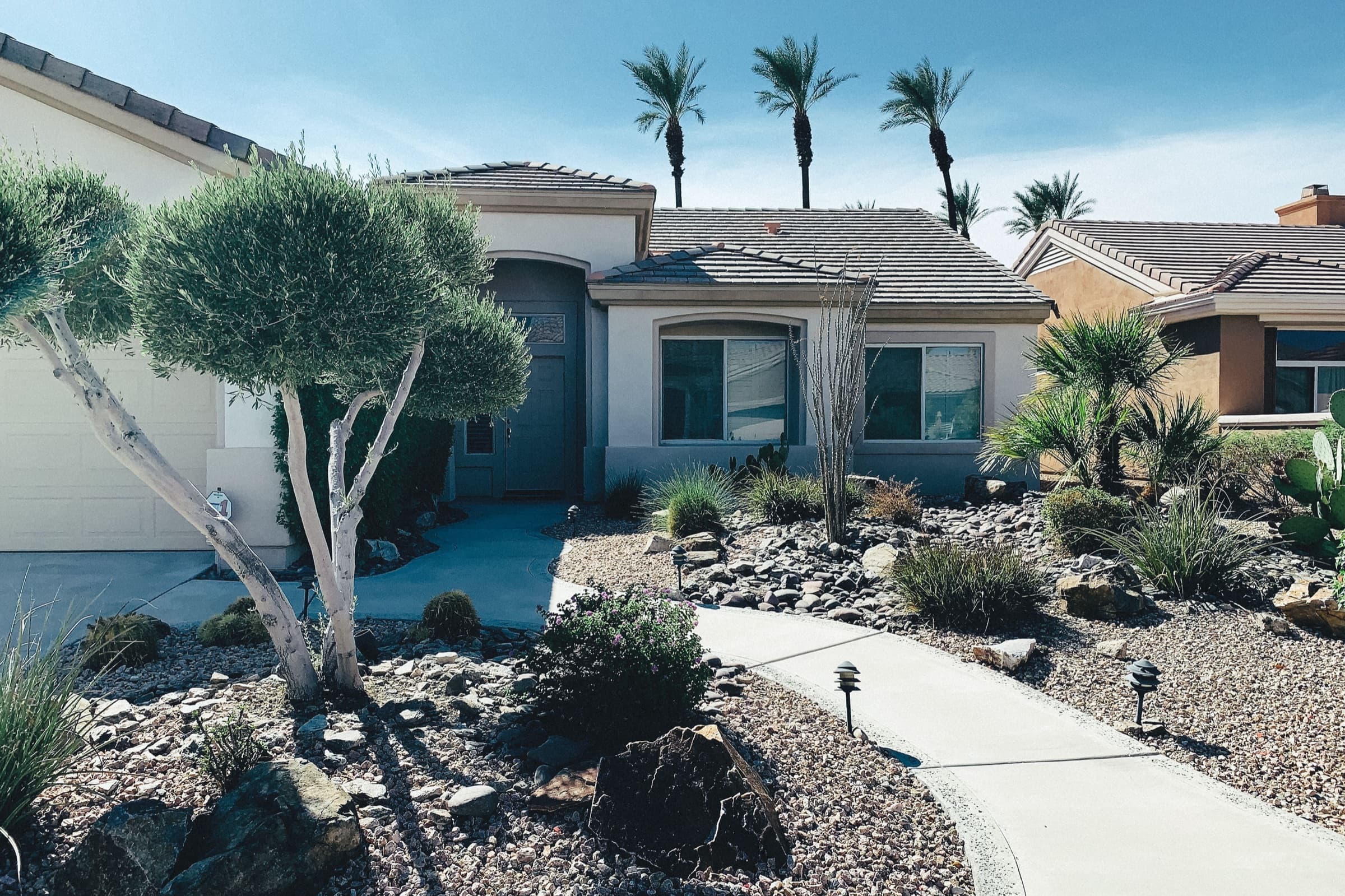 Front yard of a stucco ranch home redesigned with river rock, gravel beds, low-water plants and path lights, palms behind, the kind of setting we work with for landscape design in Harlingen, TX